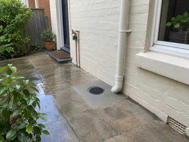 Outdoor paved courtyard with a round drain grate beside a brick wall and window in Annandale