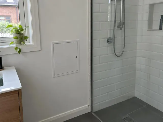 Bathroom wall with a small access panel beside a tiled shower and grey floor in Newtown